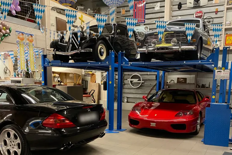 Ausstellungshalle von Koch Oldtimer: ein Citroën Traction Avant und ein Volvo Amazon auf der Hebebühne, darunter ein schwarzer Mercedes-Benz SL und ein roter Ferrari, mit blau-weisser Festdekoration